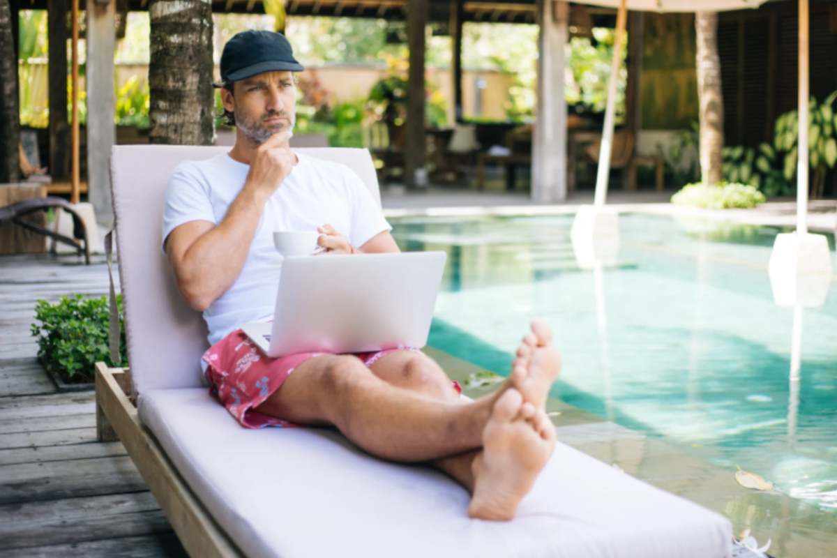 uomo steso sulla sdraio con il computer sulle gambe vicino ad una piscina